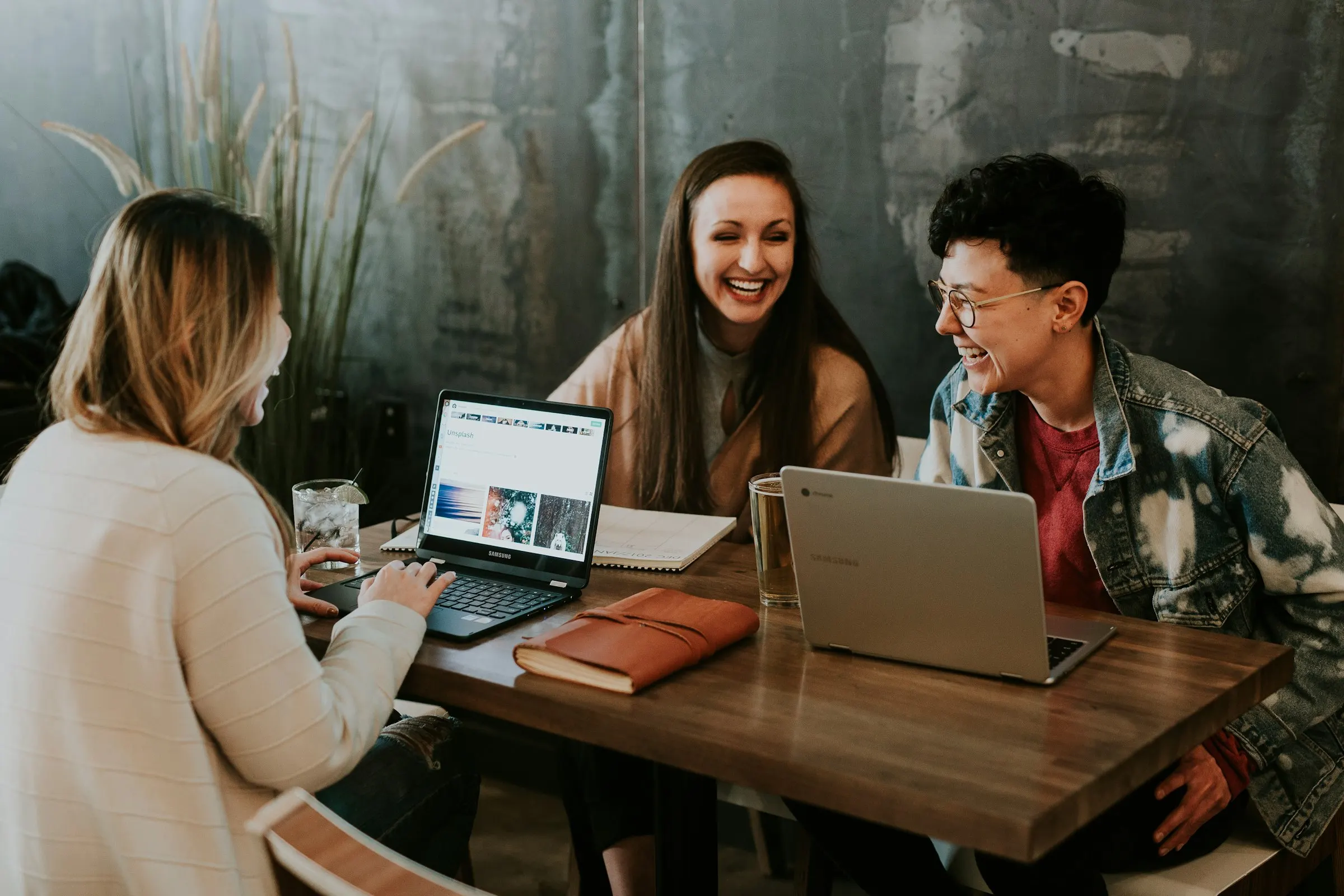 Três pessoas riem enquanto revisam um projeto no notebook, em volta de uma mesa.