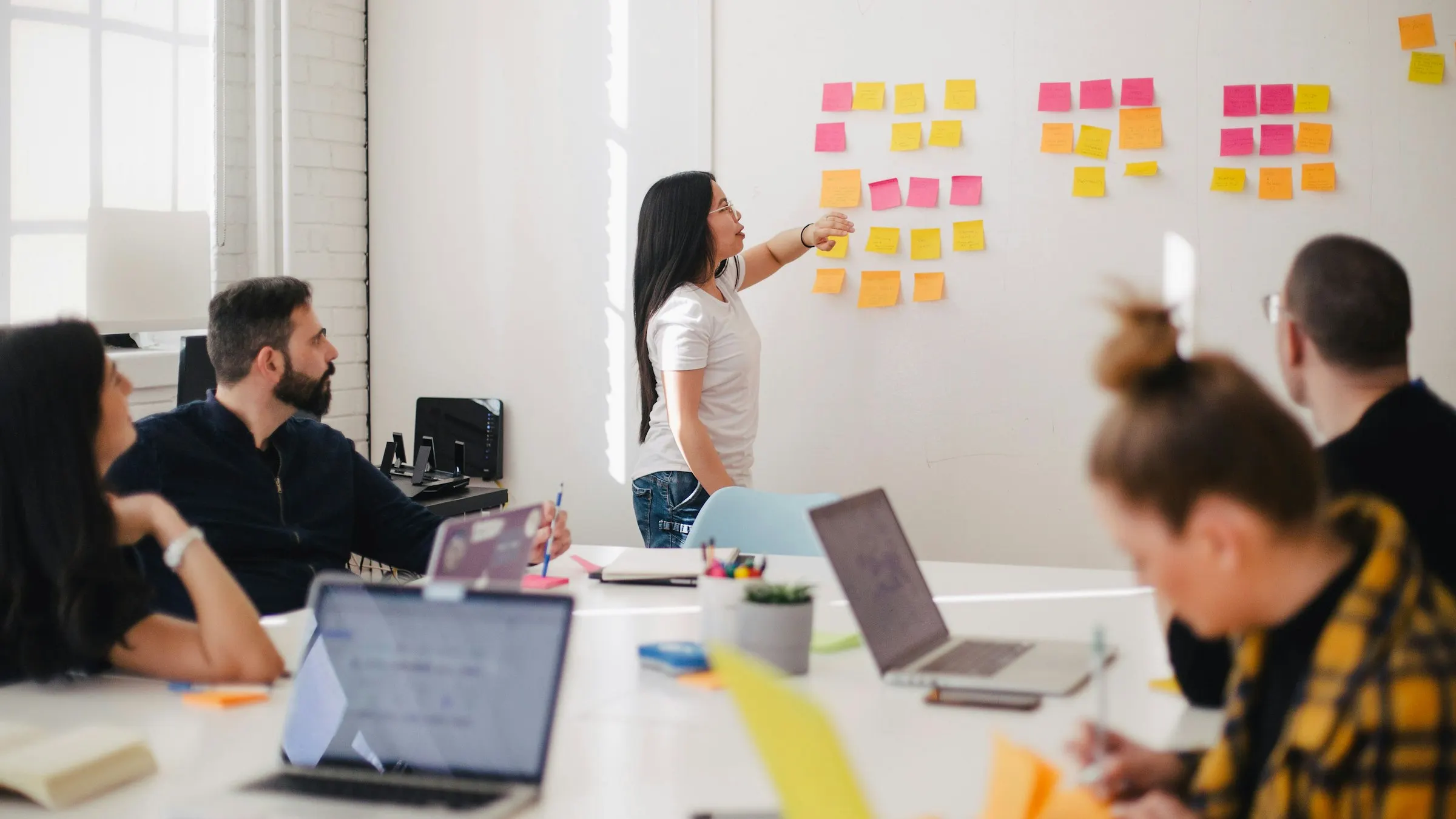 Mulher organiza notas adesivas coloridas na parede enquanto a equipe acompanha em volta da mesa.
