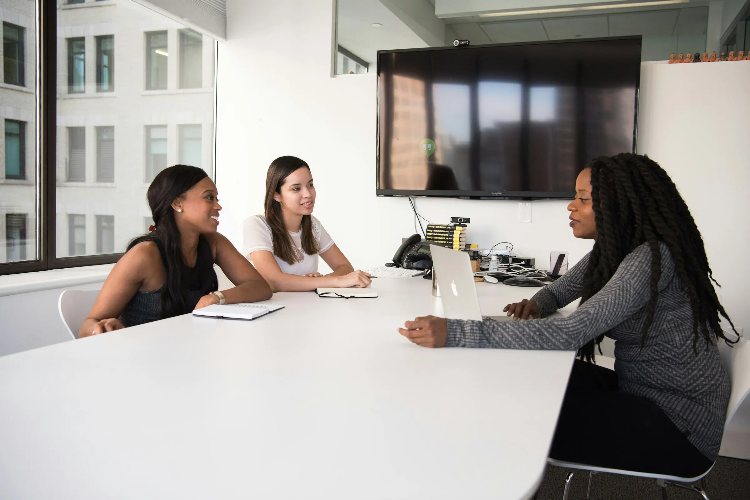 Três mulheres conversam em uma sala de reunião iluminada.