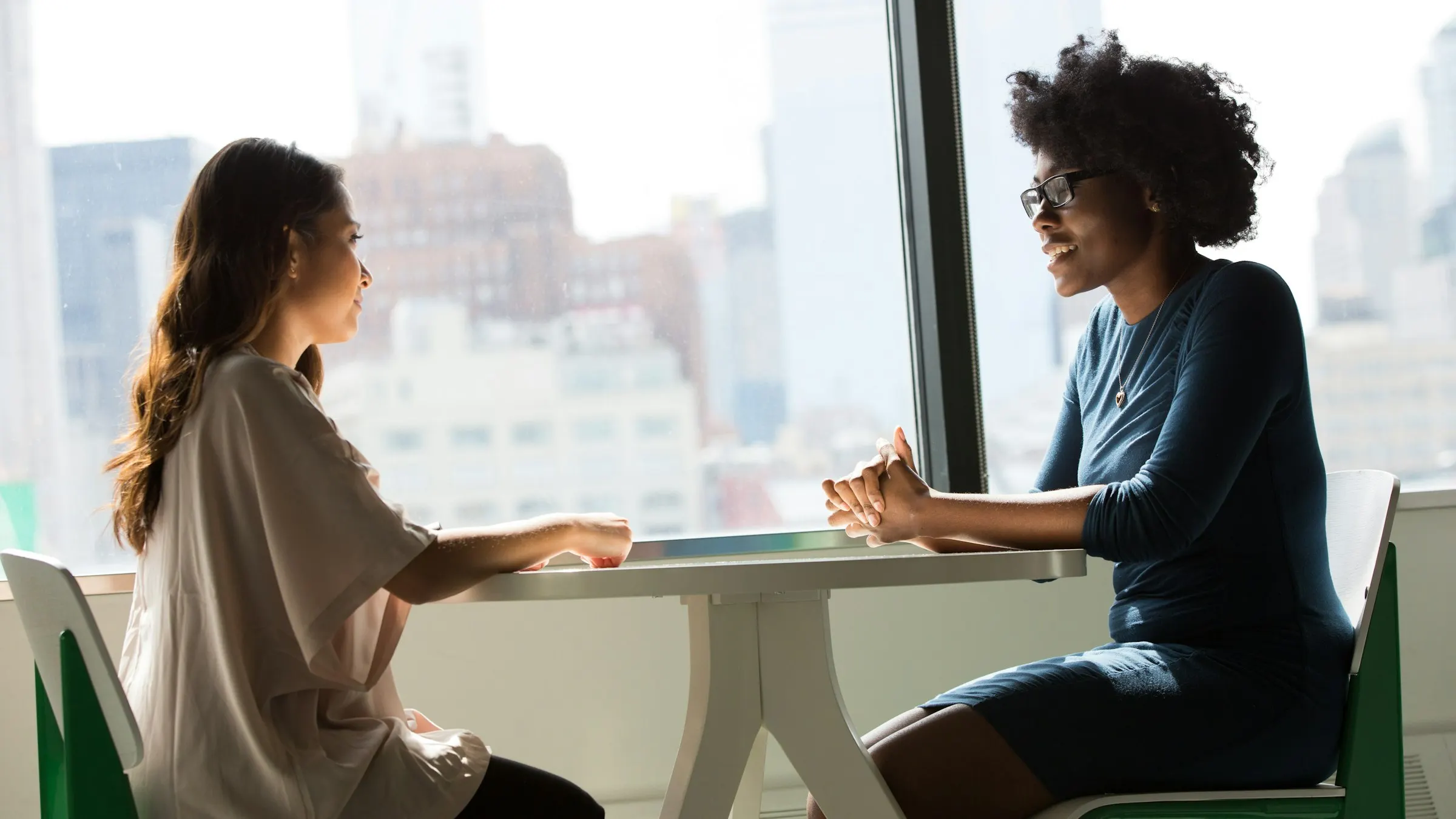 Duas mulheres conversam frente a frente em uma mesa junto à janela.
